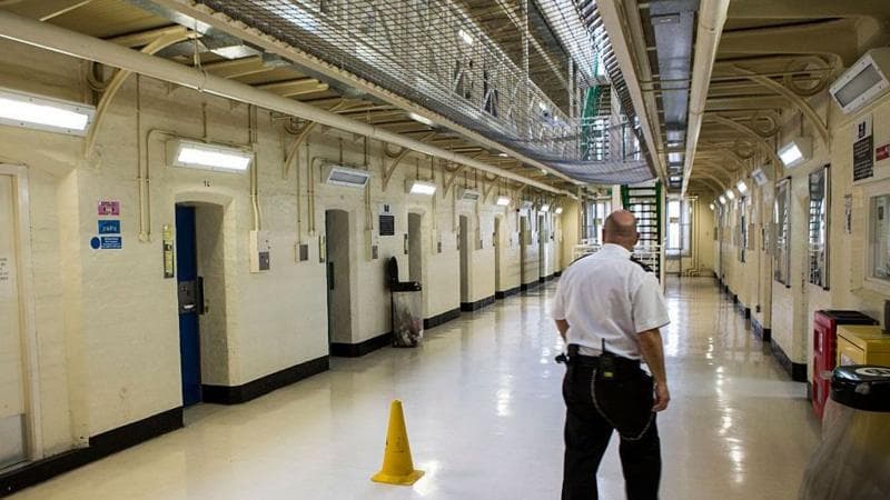 A prison guard in a white top and black trousers walks down a prison hallway. The walls and floor are white.