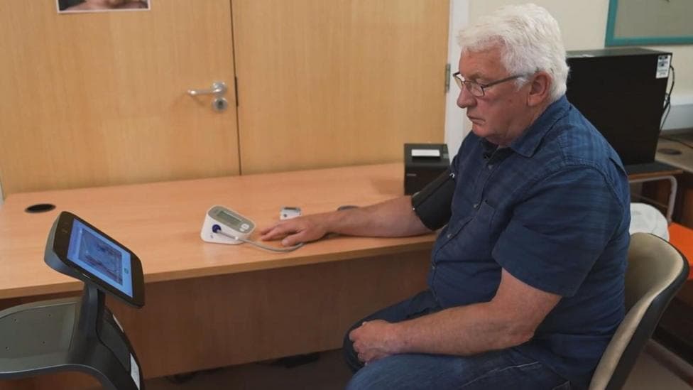 A man seated in a clinic or office consultation room using a digital health monitoring device. A blood pressure cuff is attached to one arm, with the device resting on a desk. A touchscreen unit stands nearby, and computer monitors are visible in the background.