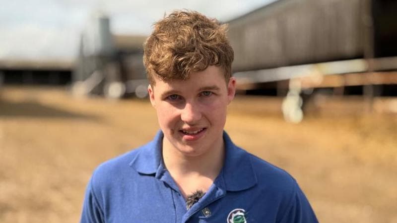 A young man is on a farm. There are sheds in the background.