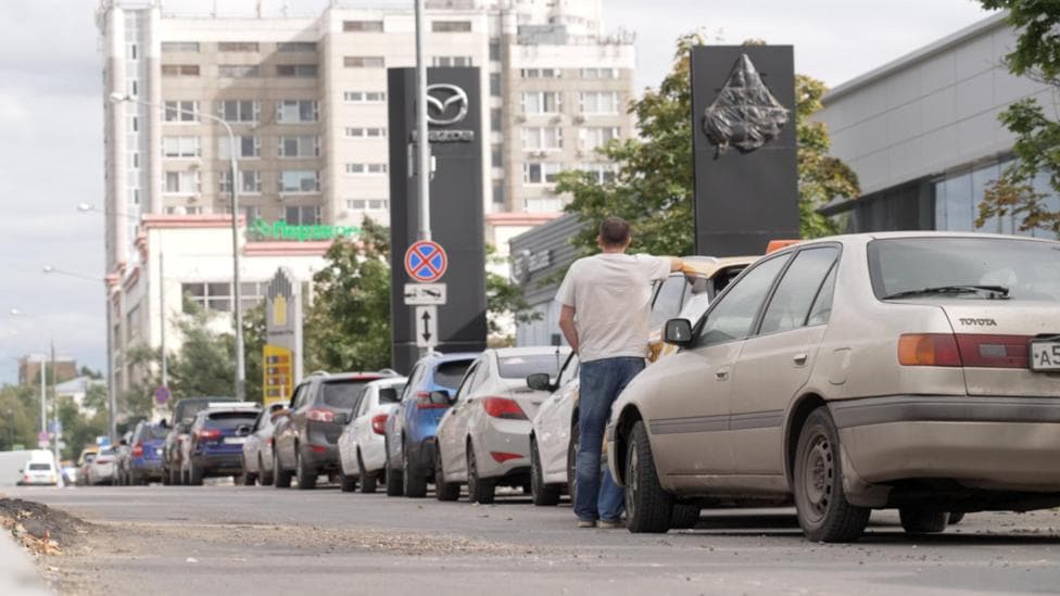 A man in a white t-shirt facing away from the camera stands with his right arm resting on his car, in a long queue of vehicles outside a petrol station in Moscow