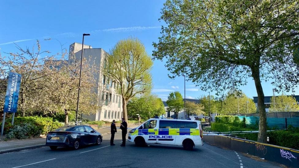 Police tape closes off the road. Two officers can be seen standing next to a police vehicle. The area is leafy with a pale building in the background.