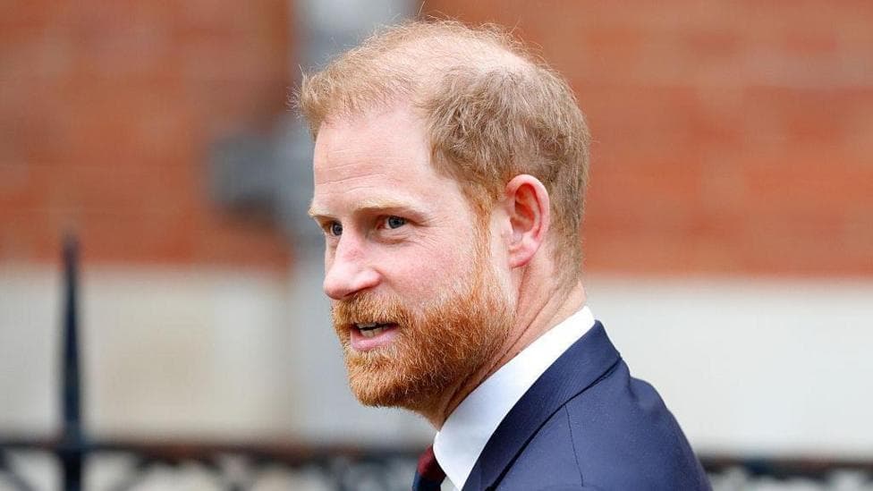 Prince Harry, a man with ginger hair and a bear, wearing a navy suit jacket with a white shirt and a tie. He is looking to the side and the background behind him is blurred.
