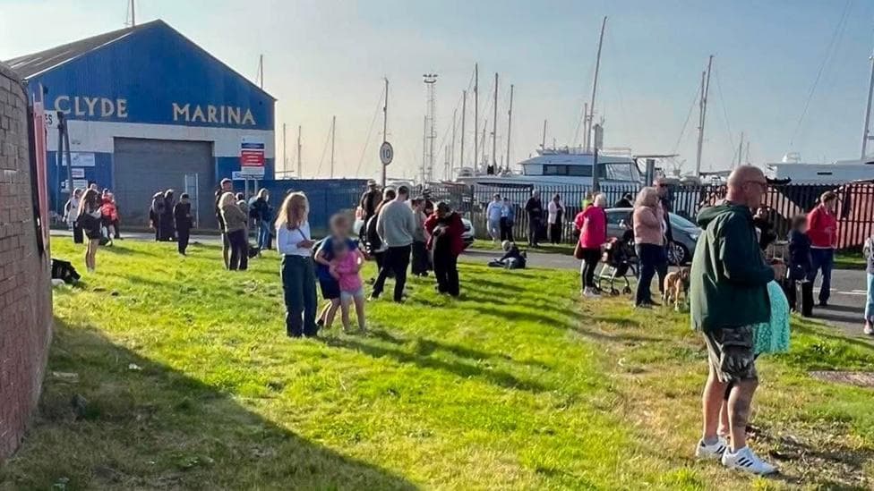 A crowd of people stands on a grassy area beside Clyde Marina, with yachts and sailboat masts visible behind a fence in the background. A large blue building marked “CLYDE MARINA” stands to the left, while people gather along the roadside and waterfront under bright sunshine. A speed limit sign and several parked vehicles are visible near the marina entrance.