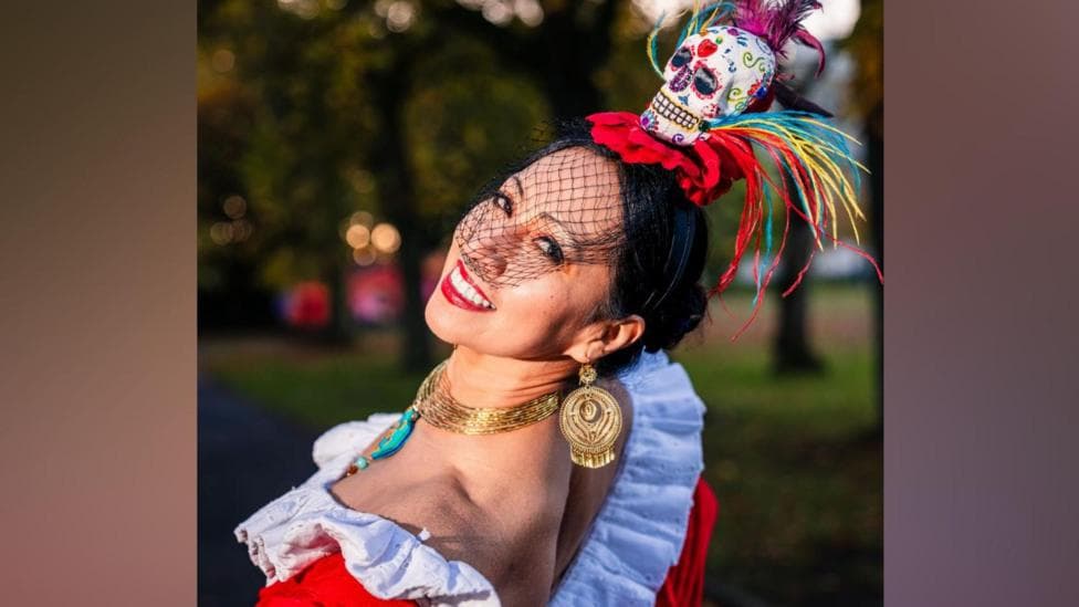 Red fascinator with skull with colourful illustrations. She is wearing an oufit with white frils, red body and a yellow/pink/yellow dress