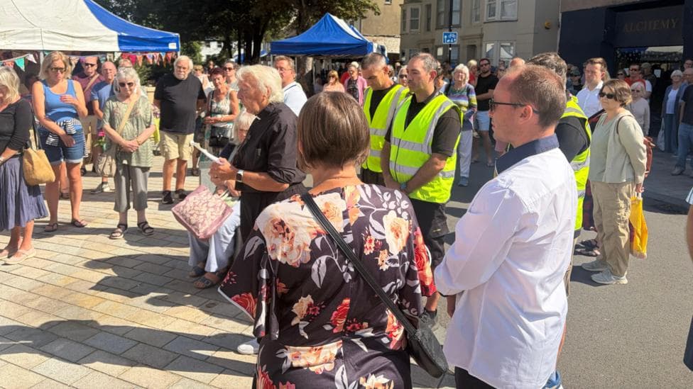 A vicar with lots of people with heads bowed in a town square daytime