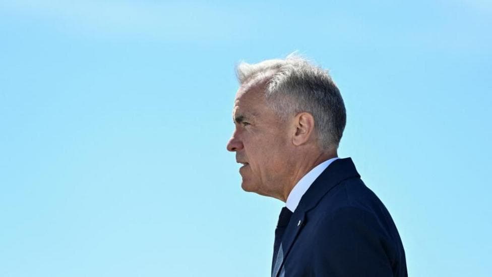 Mark Carney stands in profile, facing to the left, against a clear light blue sky. He wears a dark blue suit and tie.