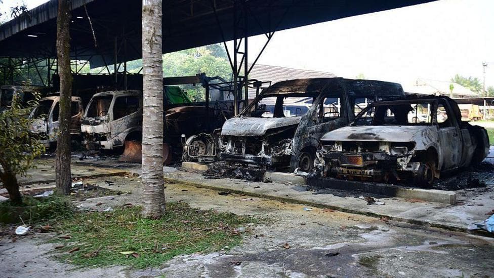 Five burned out vehicles are lined up under a tin roof structure