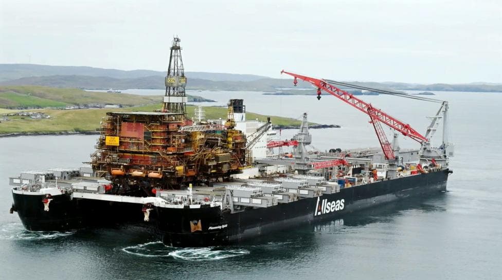 Heavy-lift vessel Pioneering Spirit carrying a large offshore oil and gas platform topside across calm coastal waters. A red lattice crane is positioned on the deck, with low-lying islands and shoreline visible in the background. The ship's hull displays the company name "Allseas."