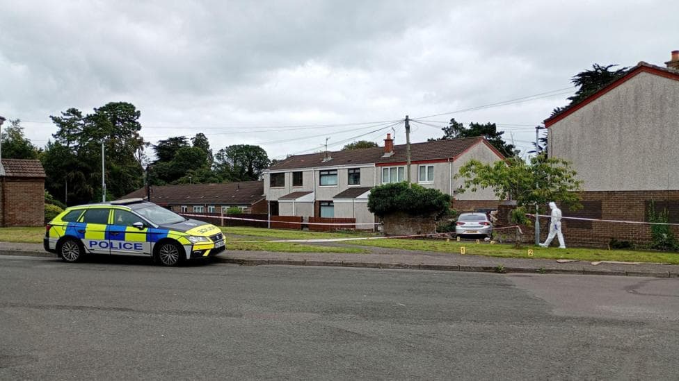 A police car parked at the side of the road in front of a row of houses. There is a forensics police officer walking along the verge and police tape.