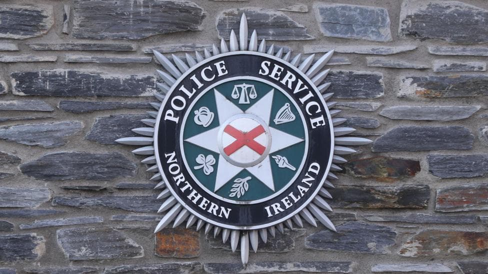 The official badge of the Police Service of Northern Ireland is displayed on a grey brisk wall.