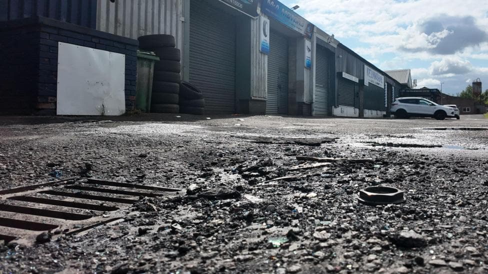 Low-angle view of an industrial estate with a row of shuttered garage units and workshops. The foreground shows a damaged asphalt surface with a drain cover, loose debris and potholes, while several parked vehicles sit beside the buildings under a partly cloudy sky.