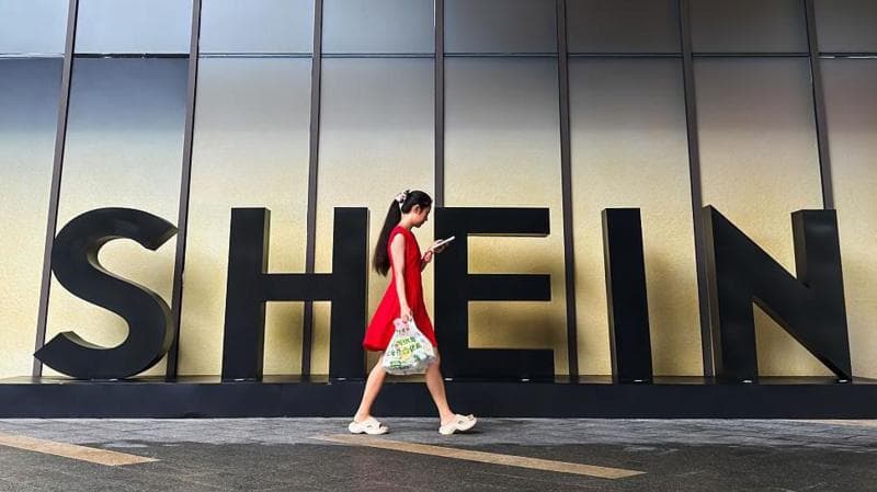 A woman in a red dress looks at her smartphone while walking past a Shein sign outside the company's office in Guangzhou, China.