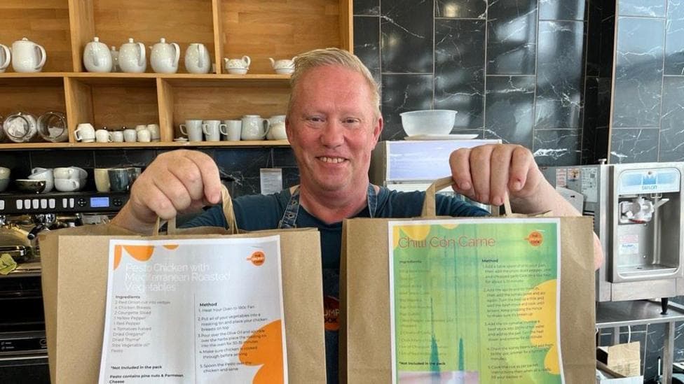 A man, Barry Ashley, stood in a cafe holding two paper bags out in front of him.