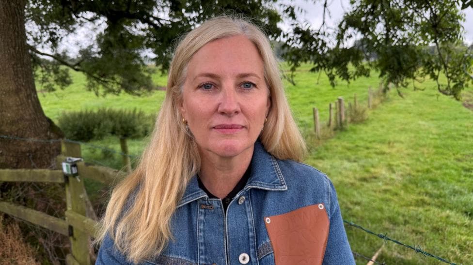 Sarah Herbison has blonde hair and a denim jacket with a brown quare on it. She is standing in front of a field with part of a tree behind her.