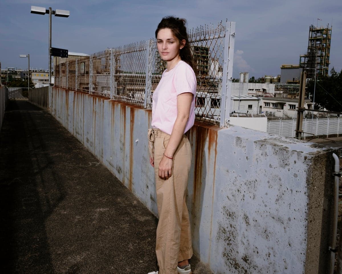 Duguy stands by a rusted wall and fence near a chemical plant