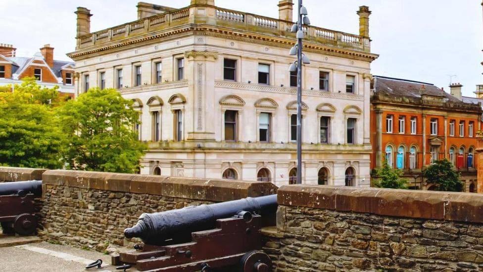 The former Danske Bank building is a large grey building.
It is viewed from Derry's historic walls were cannons are seen up against the city walls.