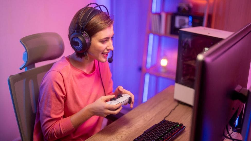 A female streamer is sitting down at a desk looking at her computer, holding a white and black PlayStation 5 controller. She is wearing a pink jumper and has a black headset on.