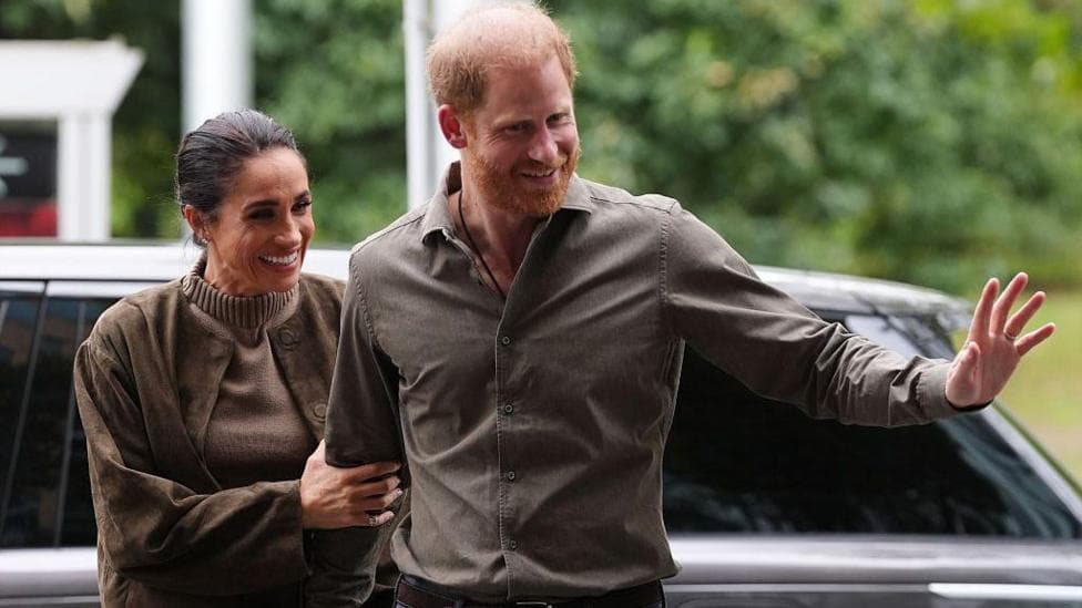 Meghan holds onto Harry's arm as they wave at crowds while they walk from a car. Both are wearing brown. They are smiling.
