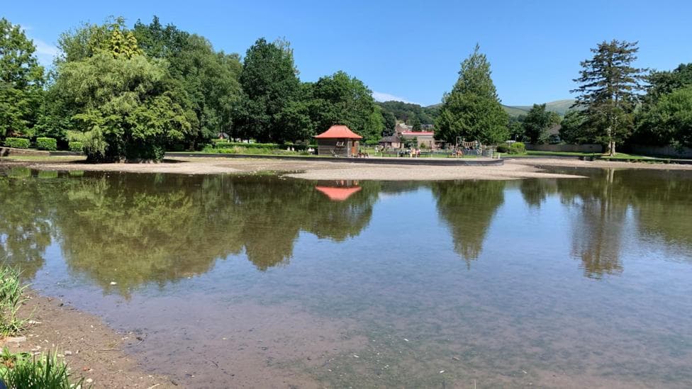 A look across Moffat boating pond with some water in the foreground but large dry patches. There are trees in the distance and the boating hut where visitors used to pay to get on the pedal boats.