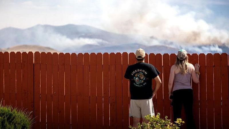 Two people look over what appears to be a domestic fence. They are looking at where smoke is rising from hills in the background.
