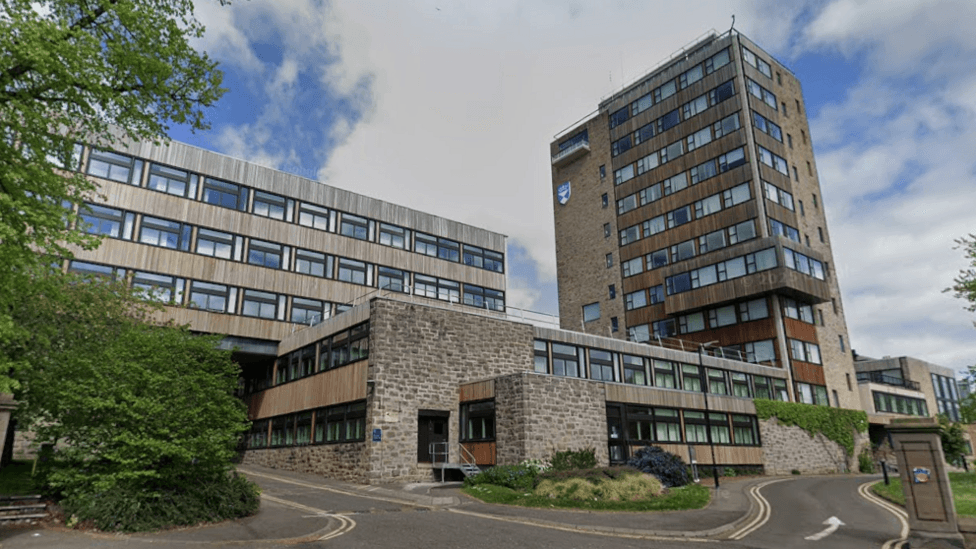 A grey/brown building at least 11 storeys high with windows. It is connected to a smaller two or three storey building of the same material on one side. On the other side connected to the smaller building is a taller building. A pathway leads up to the buildings.