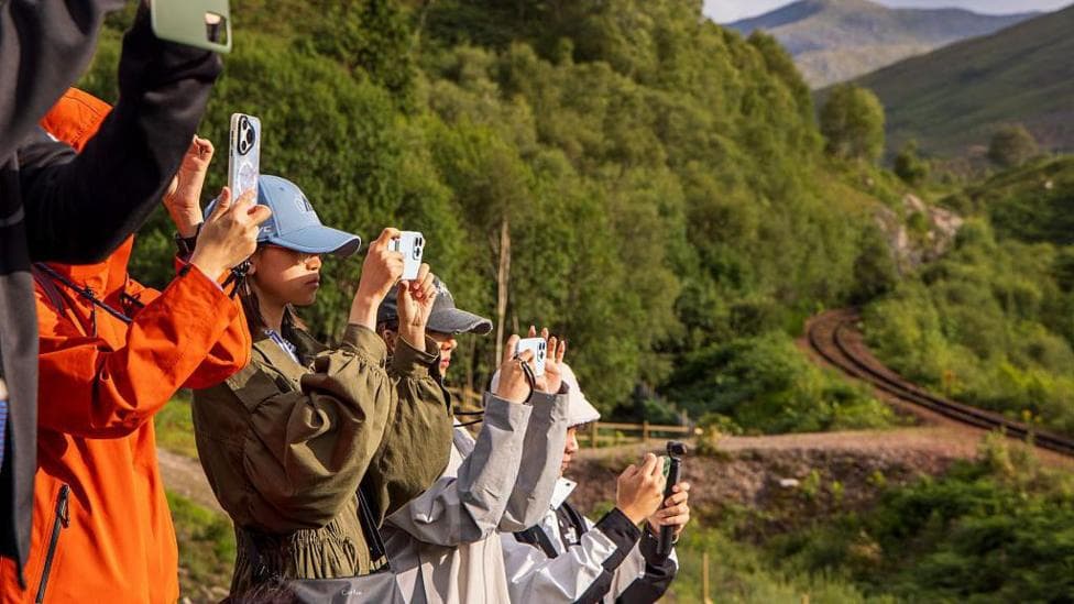 Five tourists holding up their phones to take photographs. They are in the Scottish Highlands. Mountains and a trainline can be seen in the background.