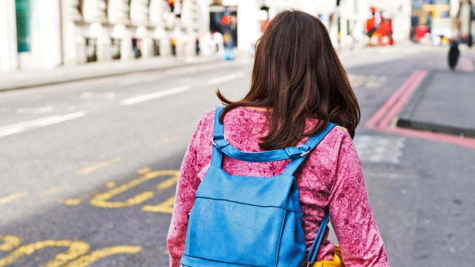 A young woman with brown hair, a pink top and a blue packpack walks down a street