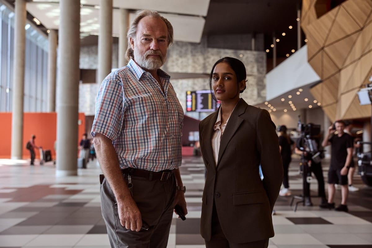 A tall man with grey hair and a grey beard, a checked shirt and grey trousers, with a slim woman with dark brown hair in a dark grey suit and pink shirt
