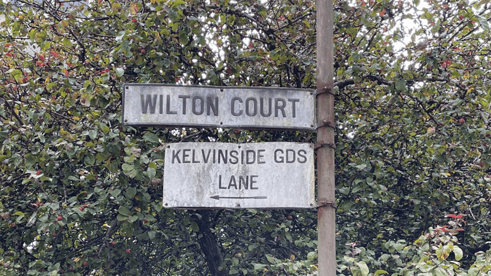 Weathered grey sign saying Wilton Court, along with a sign saying Kelvinside Gds Lane. There are trees behind it.