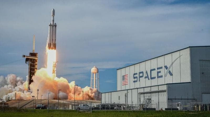 A SpaceX Falcon Heavy rocket blasting off in front of an industrial building with a huge SpaceX logo.