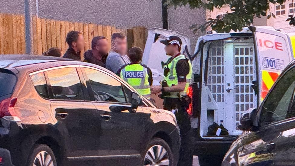 A man wearing a grey t-shirt being put into the back of a police van by three police officers