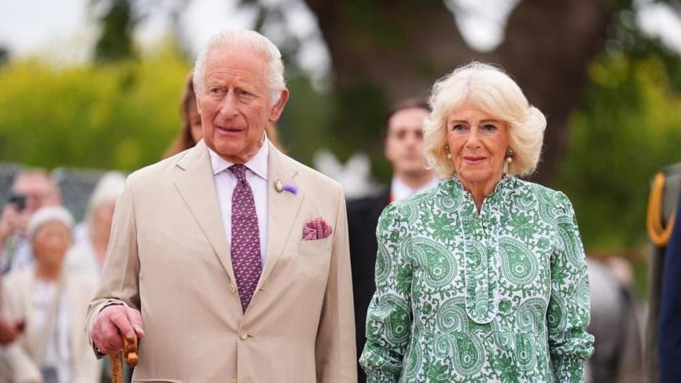 King Charles III in a tan suit and purple tie. Queen Camilla is standing next to him in a green dress.
