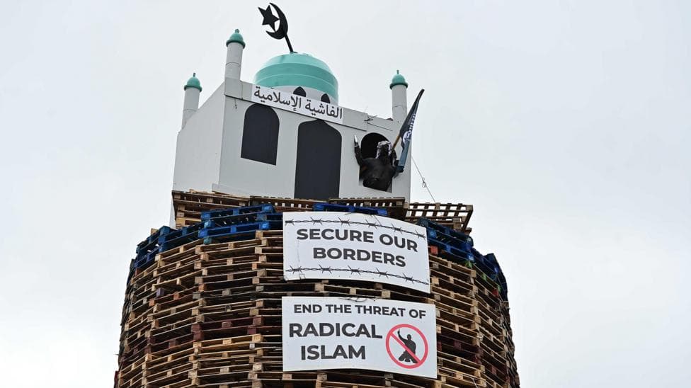 An image of the top of the bonfire, consisting of wood pallets, with a structure resembling a mosque on top. Two placards are below. One reads "secure our borders" while another reads "end the threat of radical Islam".
A figure of a man is coming out from one of the windows. The figure is wearing a headscarf and is holding a model of a knife and a flag.