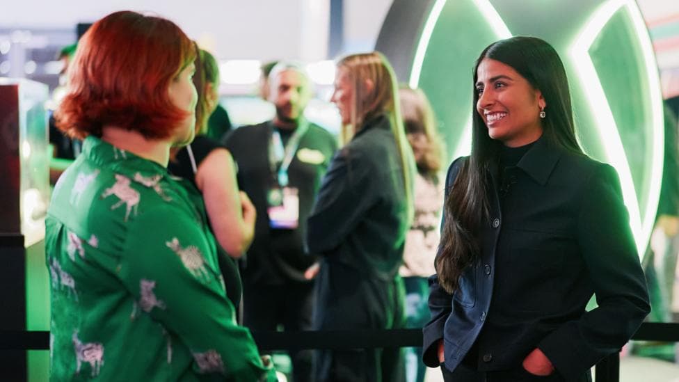 Two women stand in a convention hall with a big green Xbox logo behind them. On the left, a person with a green shirt and short red hair. On the right, a woman with long brown hair and a dark jacket.