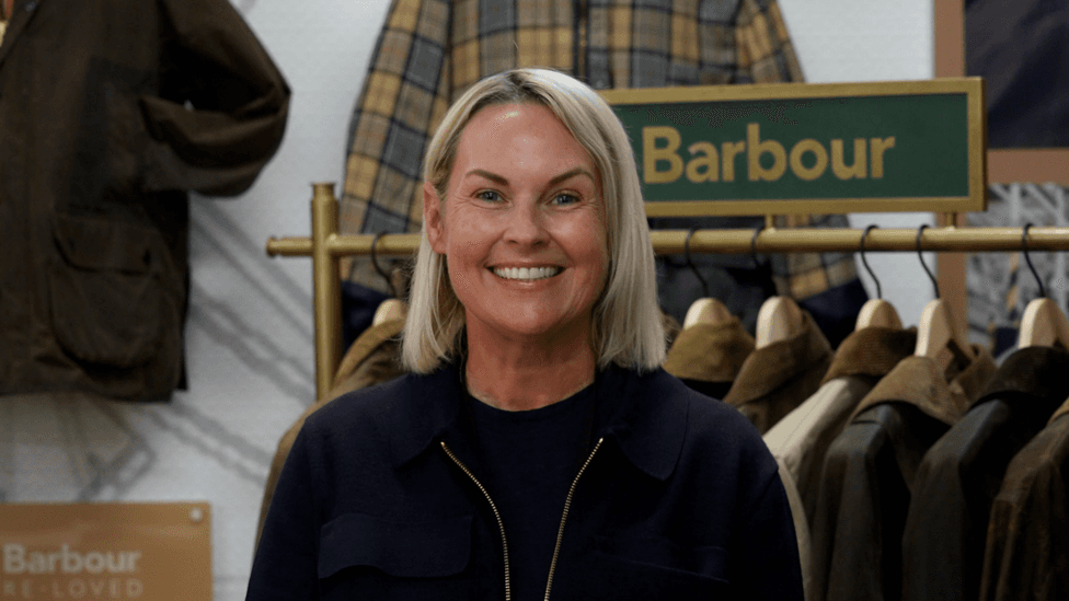 Woman standing in the foreground, wearing a dark jacket and top, facing the camera inside a clothing retail display. Behind the person is a rail of waxed jackets on wooden hangers and a green sign reading “Barbour”. Additional jackets hang on the wall, including a checked garment. A plaque on the left displays the text “Barbour Re-Loved”. The setting appears to be a branded clothing shop or exhibition area.