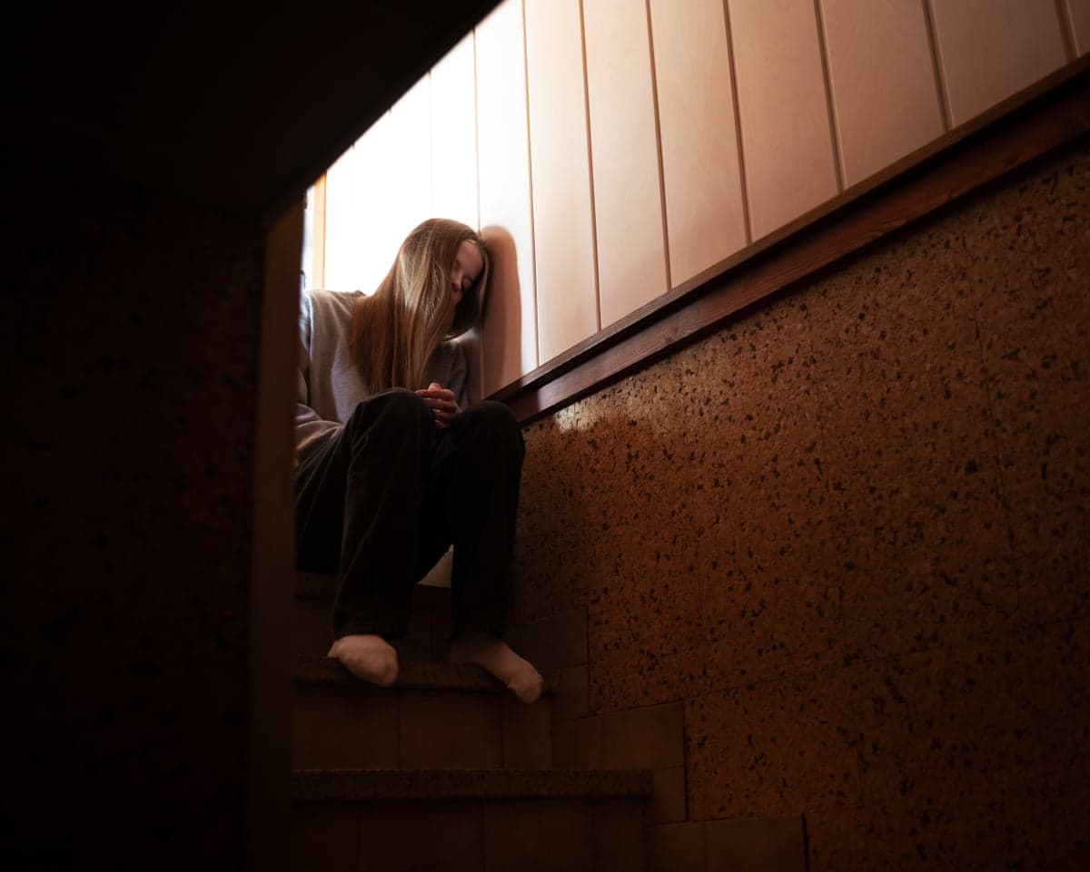 A young woman sits huddled on a staircase, head leaned against the wall, looking distressed