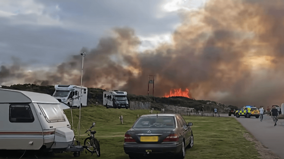 Orange flames leaping above grassland next to a campsite. There are two campervans and a caravan on the site, along with people and cars. There is also an emergency vehicle