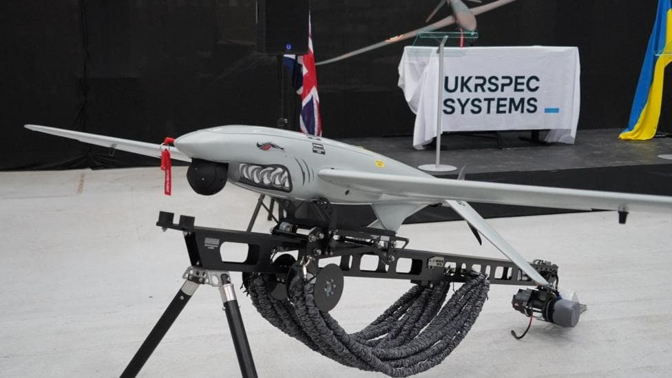 A Ukrspecsystems drone with a shark face and design on its nose propped up on a black metal stand. In the background there is a white banner which reads Ukrspecsystems, and a Ukraine flag draped next to it.
