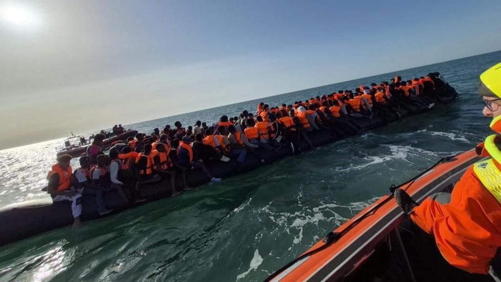 Small boat carrying a large number of people while rescue worker in a separate dinghy looks on.