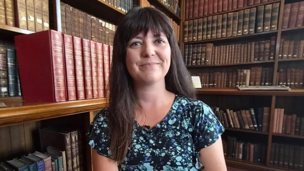 Helen Jenner is sat surrounded by books on shelves, with a large stack of red old-style books to the left of her head.