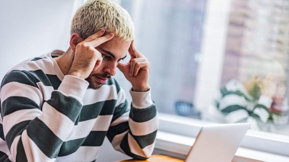 A young man with short bleached blonde hair and a beard, wearing a striped long-sleeved T-shirt, sits at a desk, looking toward an open laptop. He looks frustrated and is resting his fingers against the sides of the forehead. A large window and houseplants are visible behind him.