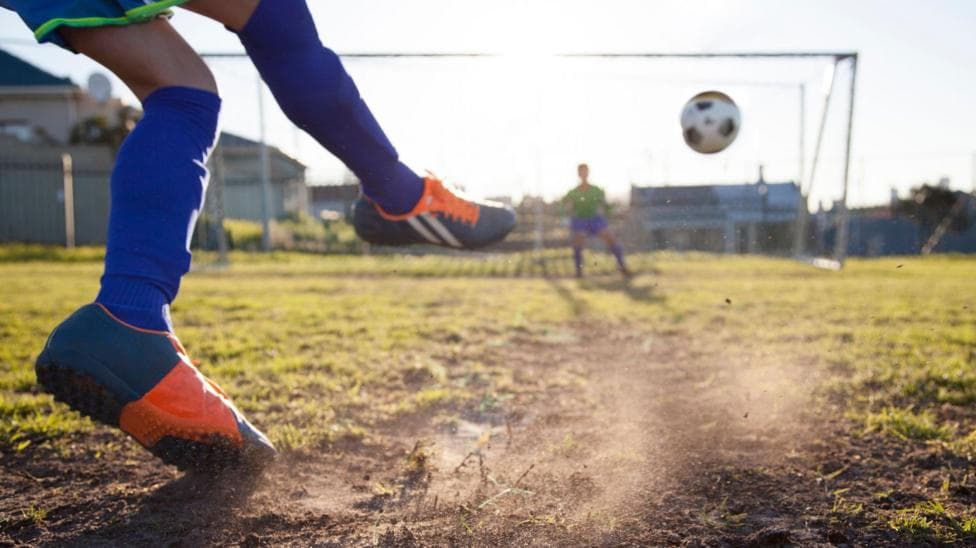 A close-up of a young boy wearing blue socks and blue and orange football boots kicking a ball into a net.