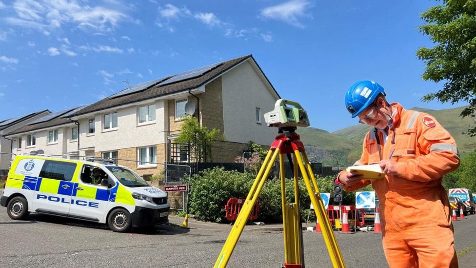 A man in orange hi vis overalls looks at a notebook, with a measuring device beside him. Police officers are visible in the background