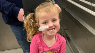 A little girl with tied-back curly blonde hair, wearing a pink t shirt with a yellow Minion character on it. She is sat in a black wheelchair with a white blanket over her legs and a feeding tube in her nose. She is smiling at the camera.