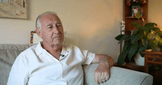 A person with short grey hair sits on a light-coloured sofa in a living room, wearing a white short-sleeved shirt and a wristwatch. In the background, a wooden shelf holds a potted plant and flowers.