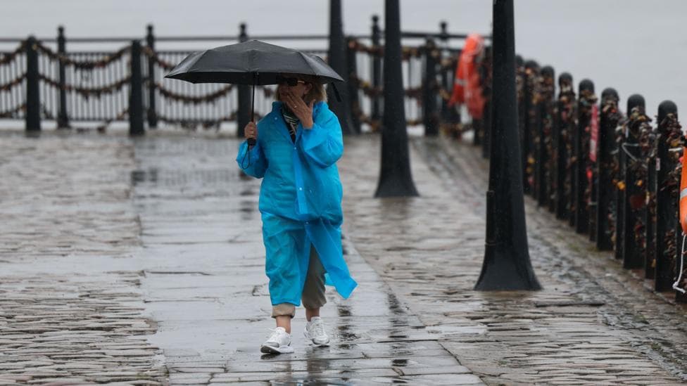 A person walks through heavy rain in Liverpool, UK in a bright blue rain coat carrying an umbrella