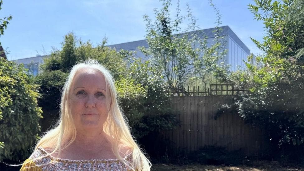 A woman with long blonde hair stands in her garden. Behind her, beyond her garden fence, is a large data centre which looks like a tall warehouse