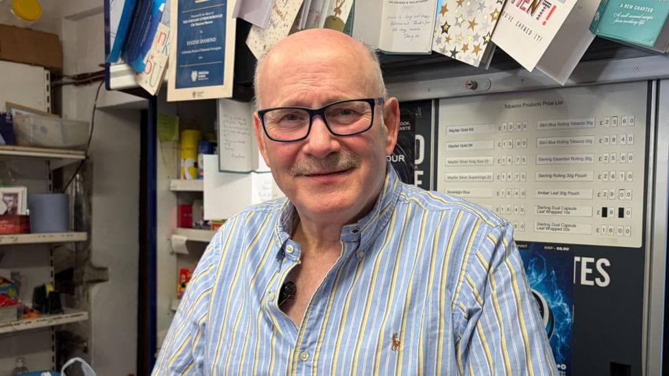 Eugene Diamond stands behind his newsagents counter. He is bald and is wearing black-rimmed glasses and a pale blue shirt with yellow stripes. Behind him is the cigarette kiosk.