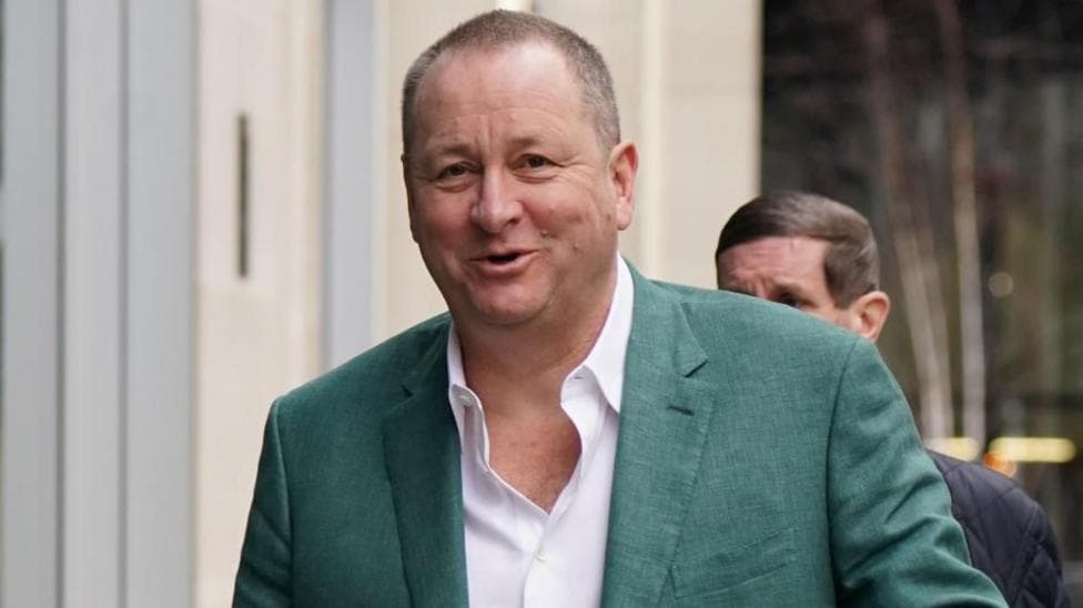 Businessman Mike Ashley wearing a white shirt, and green suit jacket, smiling at the camera as he walks with a man behind him