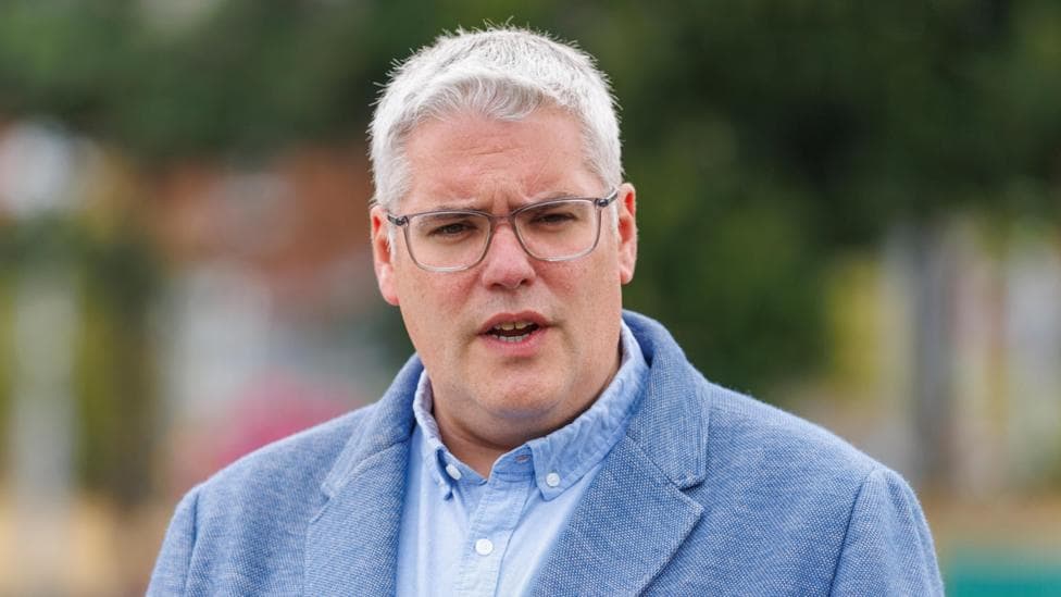 Gavin Robinson, with short grey hair, wearing glasses, a blue shirt and a blue suit jacket.